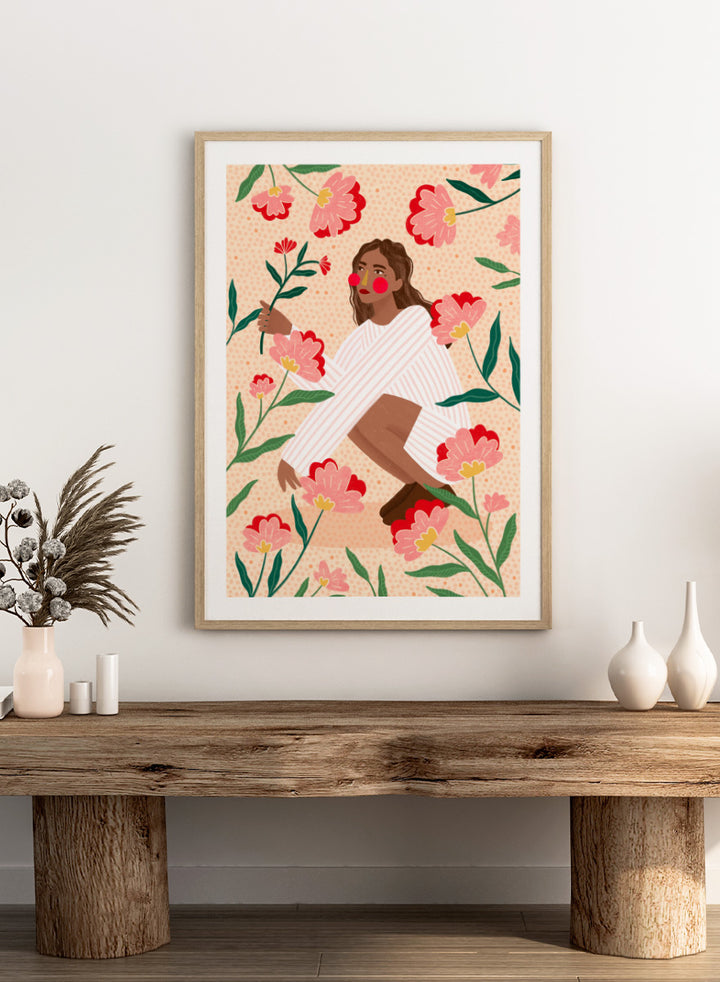 A framed art print featuring a stylized illustration of a woman in a white striped sweater surrounded by pink and red flowers on a peach background. Woman in Floral Garden by Bea Müller floral art print featuring a woman surrounded by oversized pink flowers, green leaves, and playful botanical details.