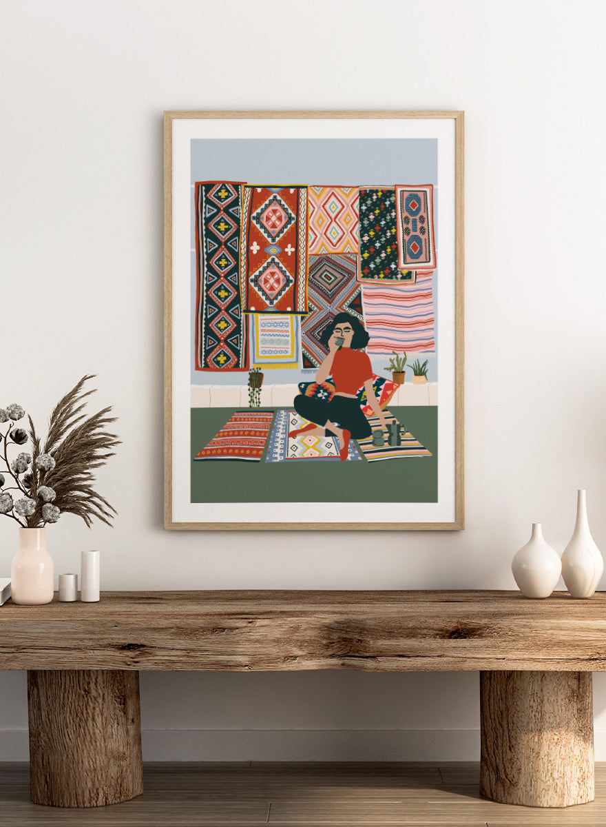 A framed art print featuring a stylized illustration of a woman sitting cross-legged on a patterned rug, surrounded by colorful geometric textiles hanging on a wall. Bohemian Coffee Morning by Ceyda Alasar interior art print featuring patterned rugs, floor cushions, potted plants, coffee, and colorful collected decor.