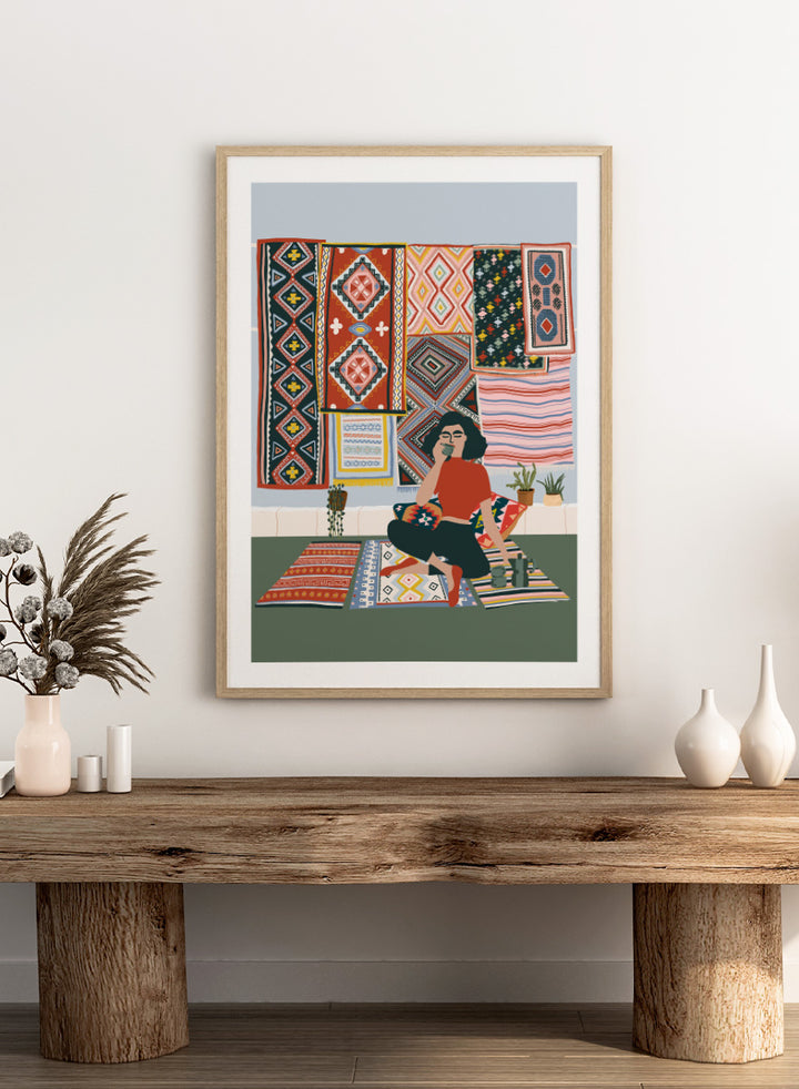 A framed art print featuring a stylized illustration of a woman sitting cross-legged on a patterned rug, surrounded by colorful geometric textiles hanging on a wall. Bohemian Coffee Morning by Ceyda Alasar interior art print featuring patterned rugs, floor cushions, potted plants, coffee, and colorful collected decor.