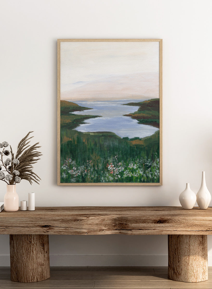 A landscape painting of a winding river or inlet between green hills, with a pale sky above and wildflowers in the foreground. Shop Wildflower Coast View by Nikita Jariwala, a coastal landscape wall art print poster featuring ocean views, green cliffs, wildflowers, and soft blue tones.