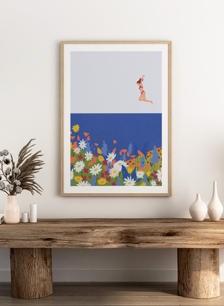 A framed art print featuring a minimalist illustration of a woman in a pink bikini jumping into a blue sea with a colorful wildflower meadow below. Wildflower Summer Jump art print by Ceyda Alasar features a joyful figure above colorful blooms, bringing playful summer energy to your walls.