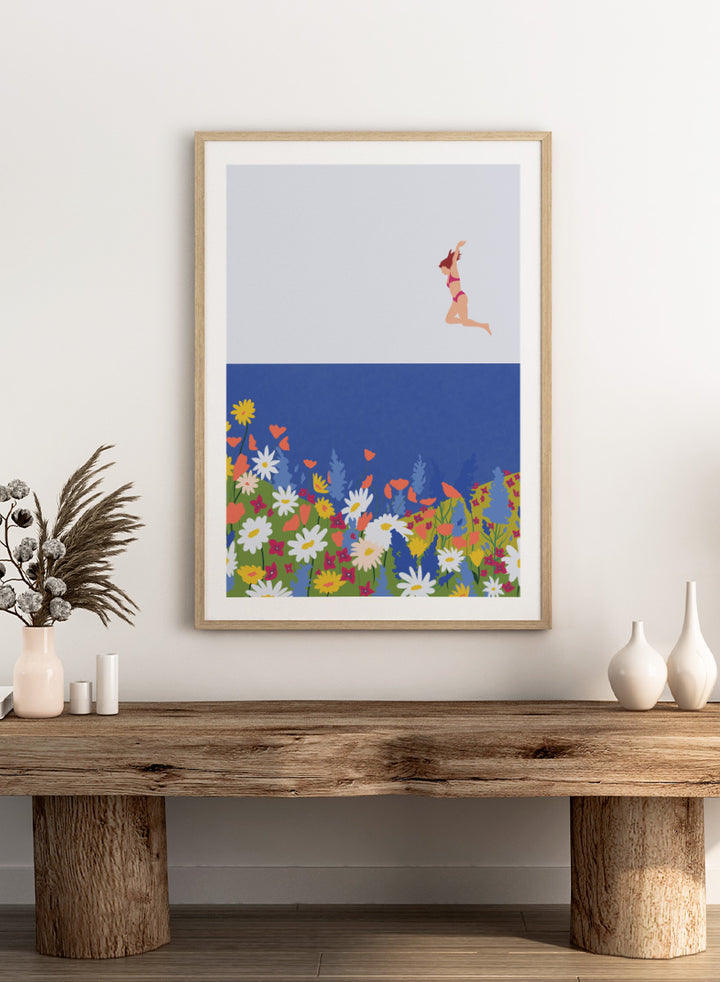 A framed art print featuring a minimalist illustration of a woman in a pink bikini jumping into a blue sea with a colorful wildflower meadow below. Wildflower Summer Jump art print by Ceyda Alasar features a joyful figure above colorful blooms, bringing playful summer energy to your walls.
