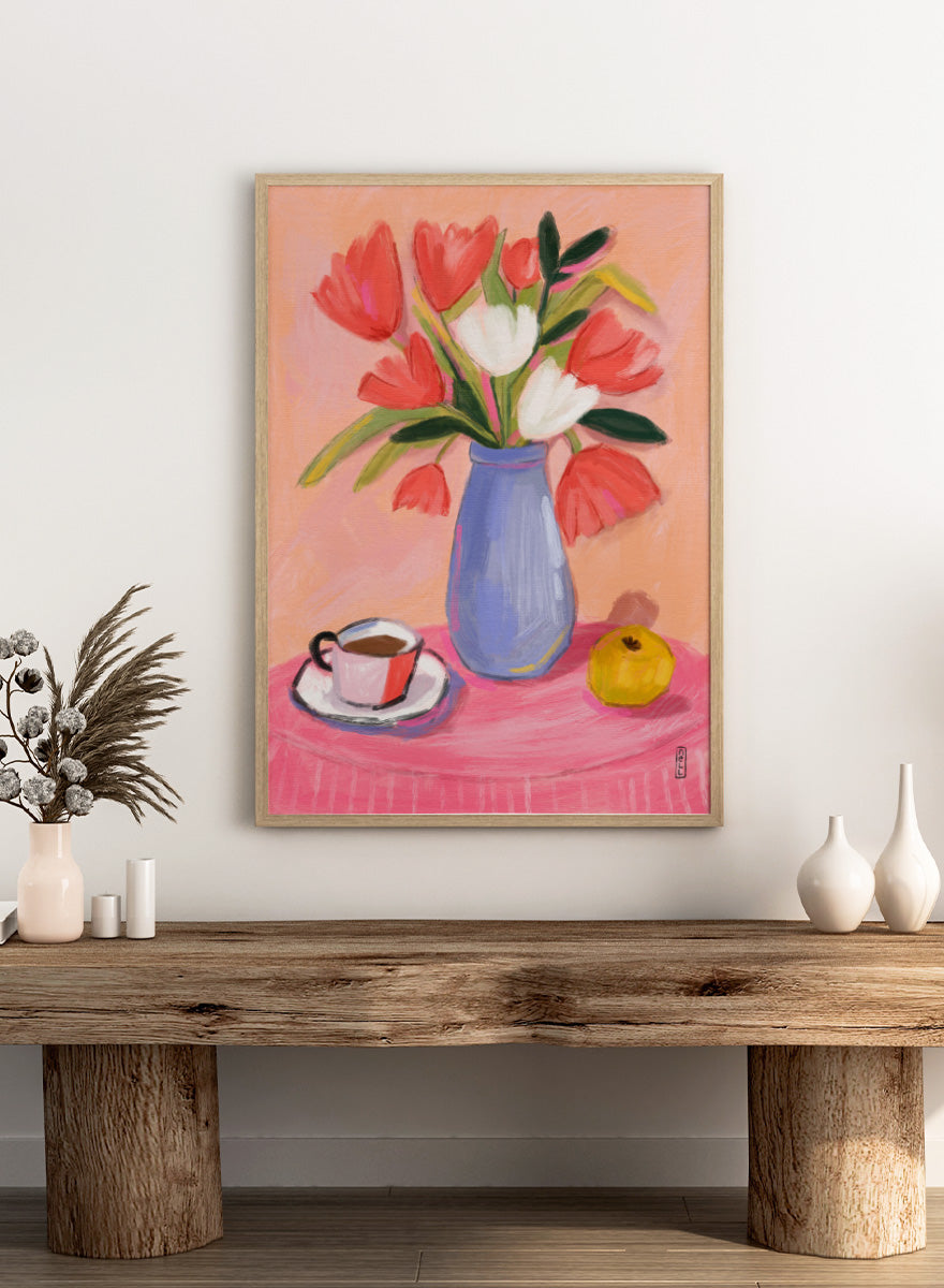 A still life painting of a bouquet of red and white tulips in a blue vase, set on a pink tablecloth next to a cup of coffee and a yellow fruit, against a peach background. Shop Fresh Tulips and Coffee by Dirkie Nell, a colorful floral still life art print featuring tulips, coffee, a blue vase, and warm pink and peach tones.