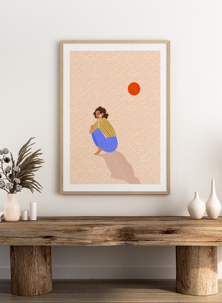 A minimalist art print featuring a stylized illustration of a person crouching in blue pants and a yellow striped top, looking up at a small orange sun against a light beige dotted background, framed with a white mat and black border. Pensive Woman Under Sun by Bea Müller art print featuring a seated woman beneath an orange sun in bold blue, mustard, and soft neutral tones.