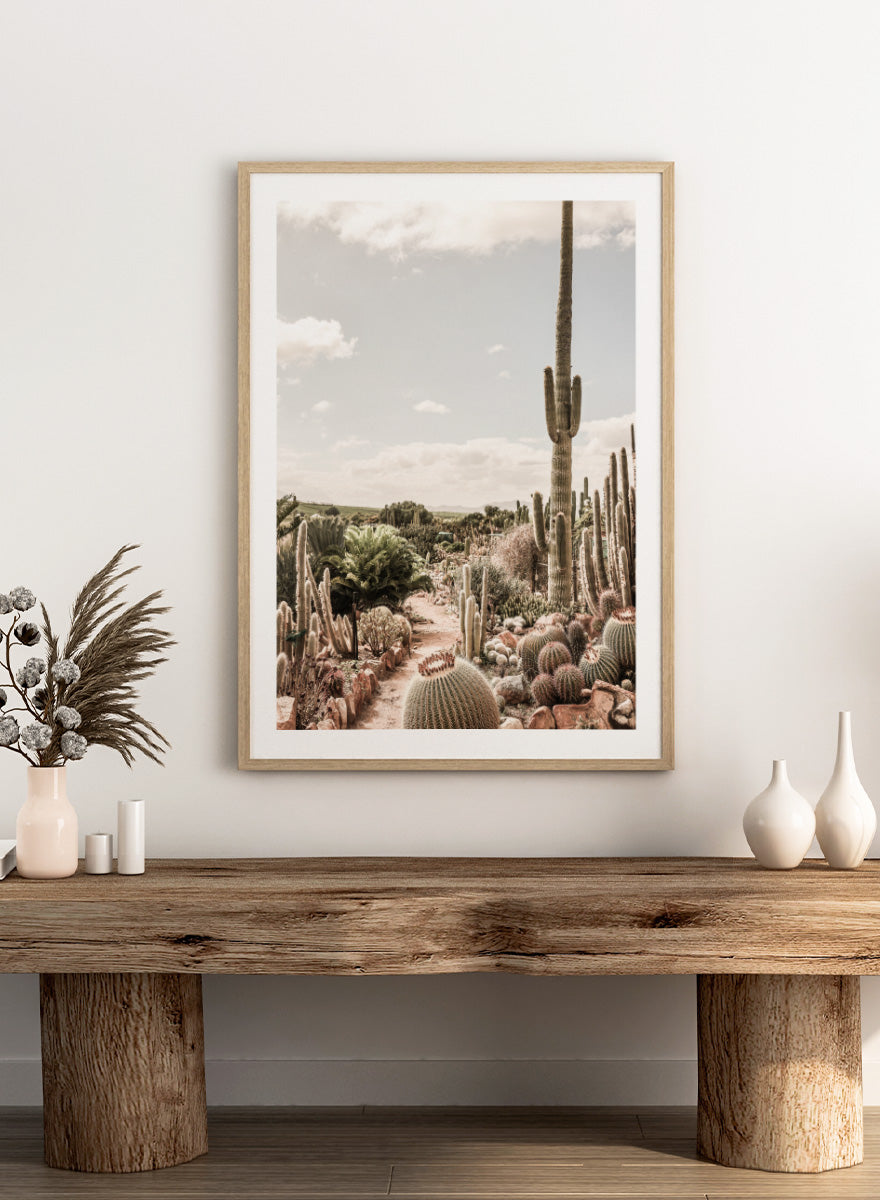 A framed art print depicting a desert landscape with various cacti and a tall saguaro under a cloudy sky. Cactus Farm by Shot by Clint desert photography print featuring towering cacti, winding garden paths, warm stone, and layered botanical texture.