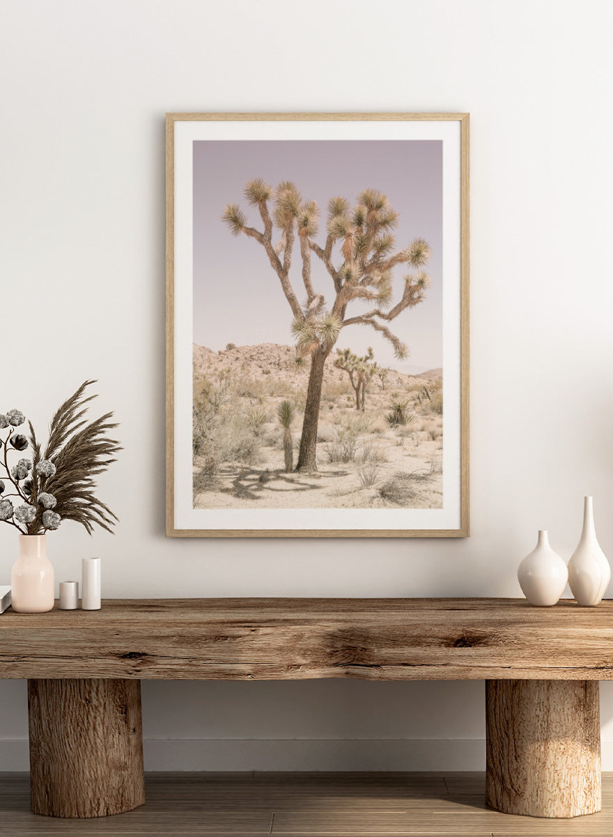 A framed art print depicting a desert landscape with a prominent Joshua tree in the foreground, set against a pale lavender sky and sandy hills. Desert Serenity by Shot by Clint features a Joshua tree desert landscape in soft earthy tones with a pale sky and calm, sun-washed atmosphere.
