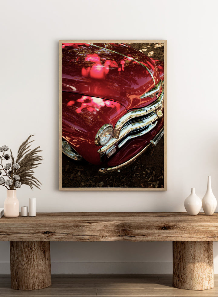 Close-up of a vintage red Chevrolet car body with chrome grille and reflections of trees on the glossy paint, photographed outdoors on mulch. Vintage Red Car by Shot by Clint automotive photography print featuring a classic Chevrolet, deep red paint, polished chrome, and sculptural vintage details.