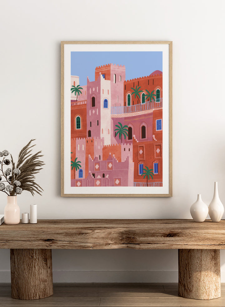 A framed art print featuring a stylized illustration of Moroccan-style buildings in shades of terracotta, pink, and cream, accented with green palm trees and blue window details, set against a light blue sky. Vibrant Moroccan City by Ceyda Alasar travel art print featuring layered terracotta architecture, arched windows, palms, and colorful Moroccan details.