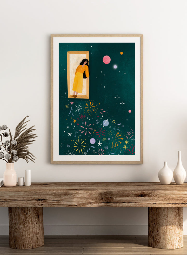 A framed art print featuring a whimsical illustration of a woman in a yellow dress standing in a mirror, with her reflection spreading colorful fireworks and space-themed motifs like planets and stars across a dark teal background. Shop Girl and Cosmic Fireworks by Bea Müller, a figurative wall art print poster featuring a woman, planets, stars, and colorful cosmic fireworks in deep green tones.