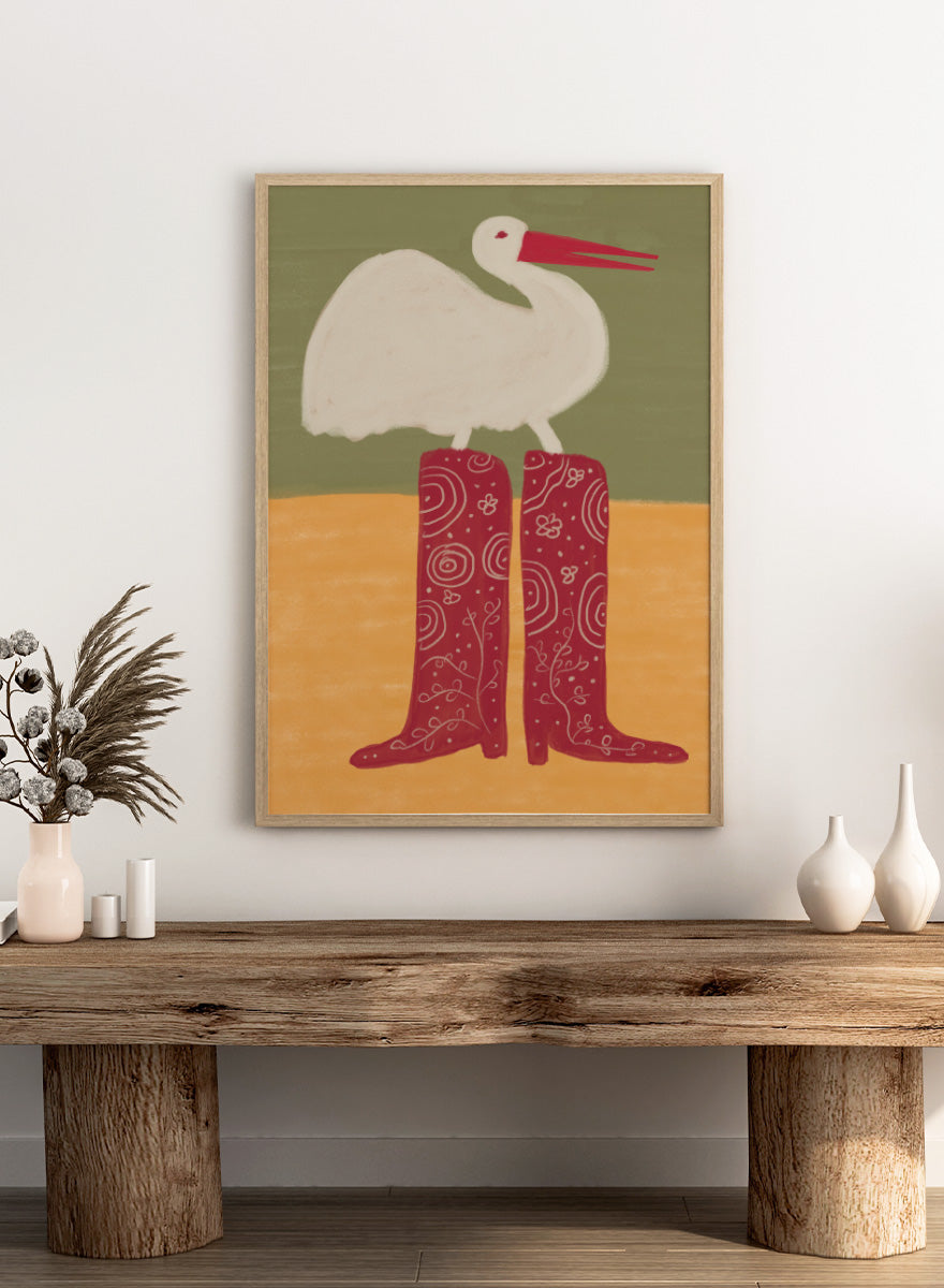 A stylized art print featuring a white stork with a red beak standing on a pair of tall red boots with white swirl patterns, set against a muted green and ochre background. Bird in Red Boots by The Art Concept playful animal art print featuring a white bird in oversized red patterned boots with soft green and ochre tones.