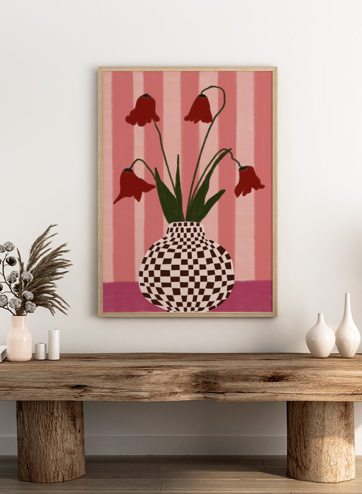 A stylized floral artwork showing four dark red bell-shaped flowers with long green stems rising from a black-and-white checkerboard vase, set against pink vertical stripes and a pink base. Whimsical Flowers Checkered Vase by The Art Concept still life art print featuring red flowers, a checkered vase, and a soft pink striped background.