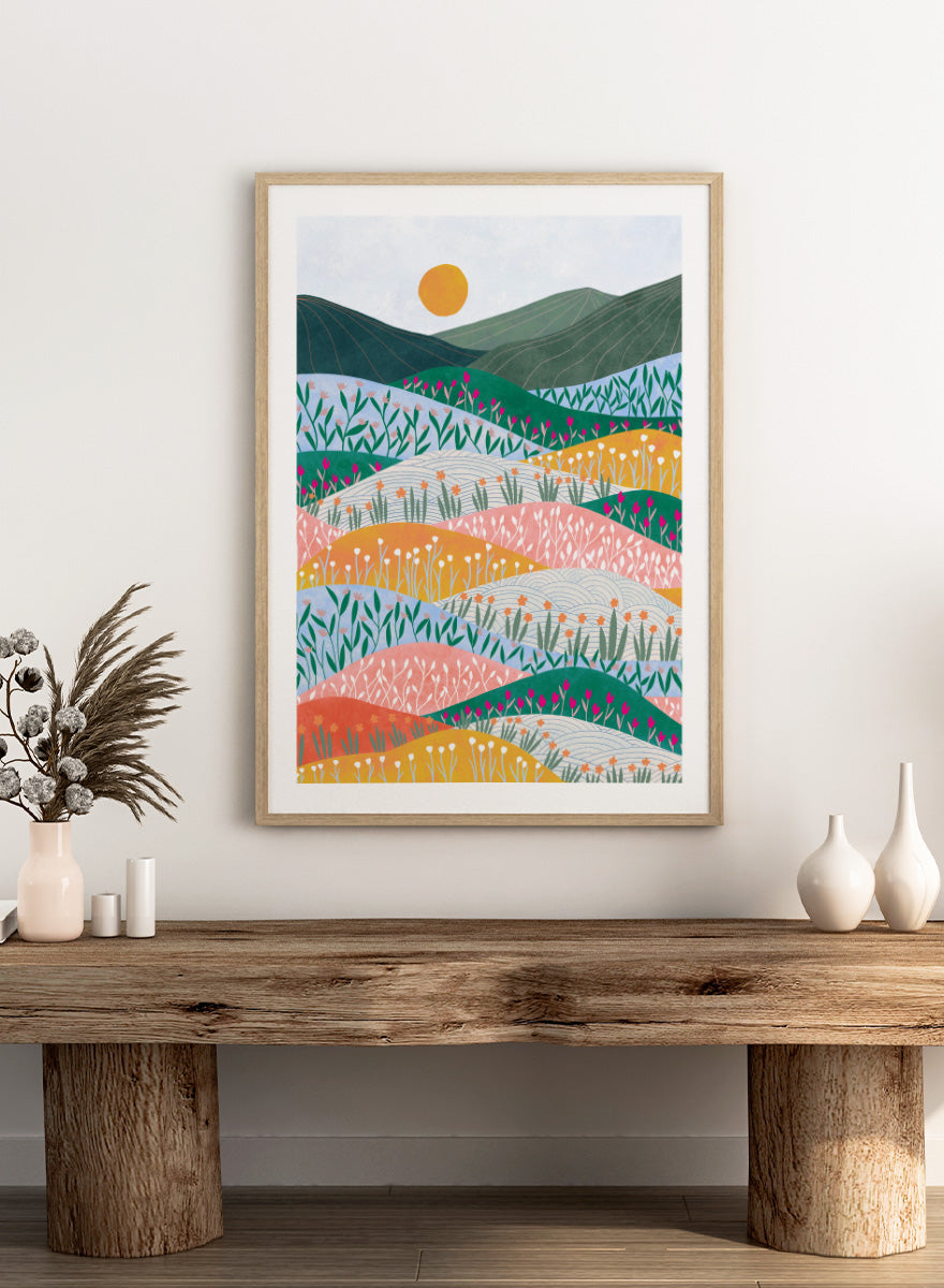 A framed art print featuring a stylized landscape illustration with rolling hills, wildflowers, mountains, and a sun in pastel and bright colors. Colorful Flower Hills by Ceyda Alasar botanical landscape art print featuring layered wildflower fields, rolling hills, mountains, and a golden sun.