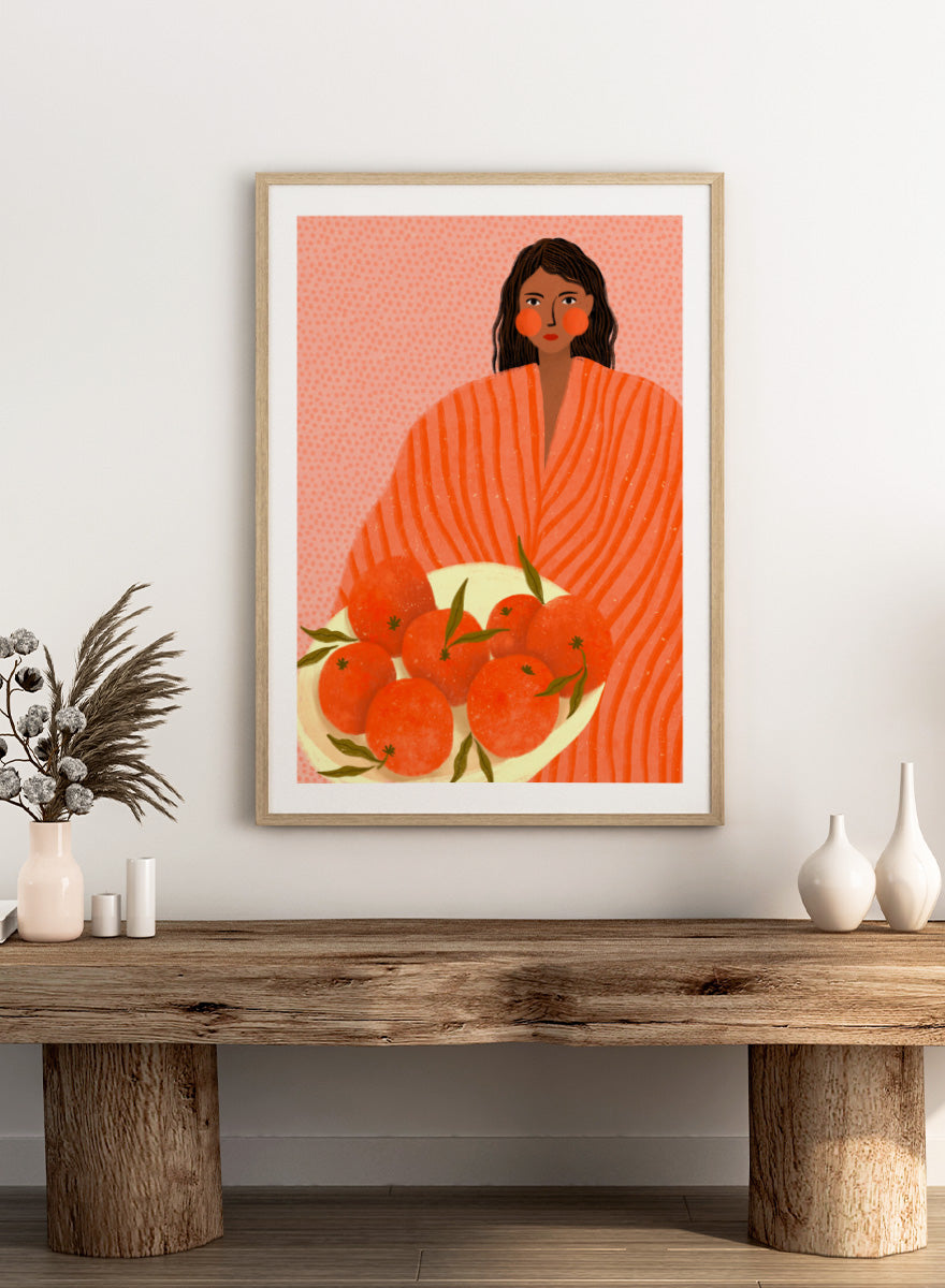 A stylized art print featuring a woman with dark hair wrapped in a bright orange striped blanket, holding a white plate of oranges against a pink dotted background. Woman Holding Oranges by Bea Müller colorful kitchen art print featuring a woman carrying fresh citrus fruit in warm orange and coral tones.