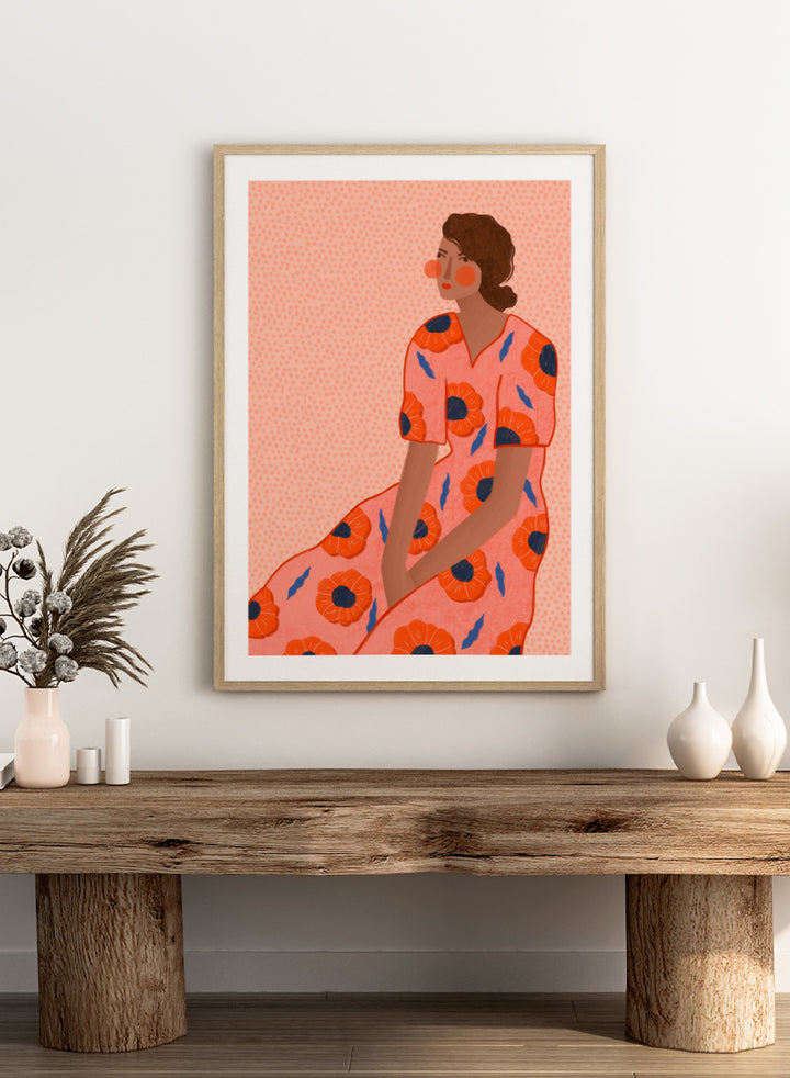 A stylized fashion illustration of a seated woman in a coral pink dress with large orange poppy motifs and blue leaf accents, shown with a white border and black frame. Stylish Poppy Portrait by Bea Müller floral woman art print featuring a pink dress covered in orange poppies and blue leaves for colorful modern wall decor.