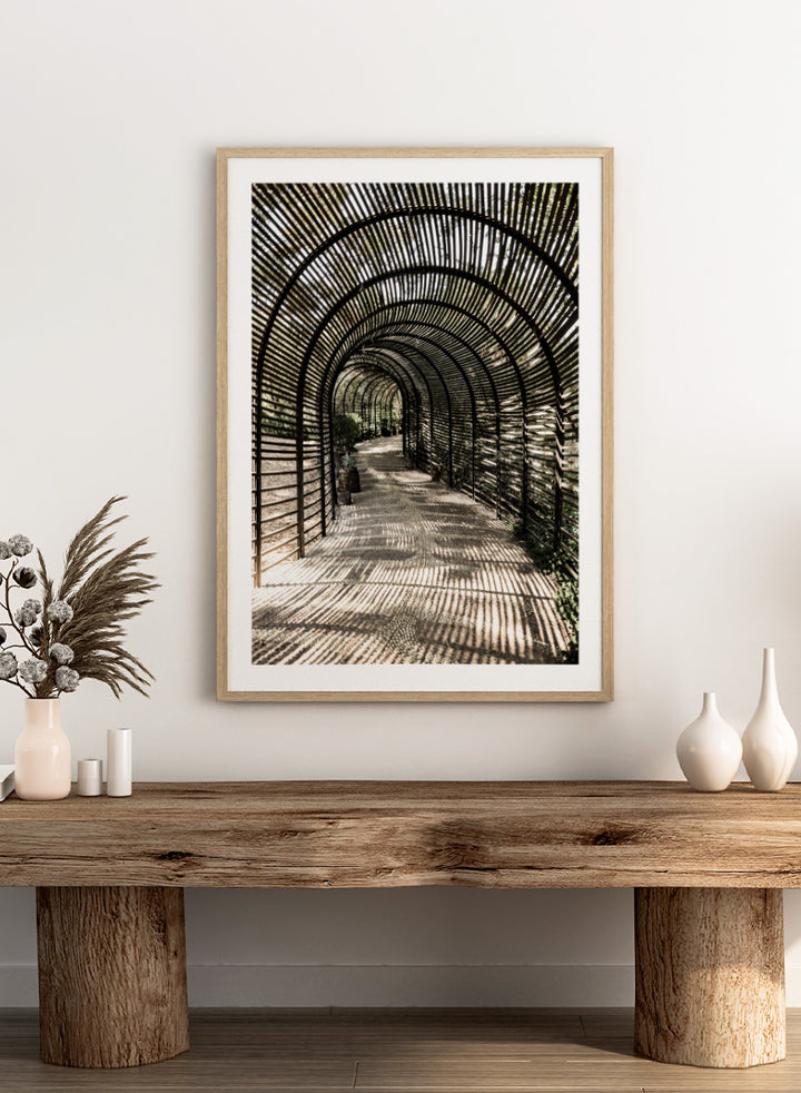 A framed art print featuring a perspective view of a garden pathway enclosed in a dark arched trellis structure, casting striped shadows on the ground. Shadow Garden by Shot by Clint architectural photography print featuring a curved wooden walkway, striped shadows, garden plants, and textured natural light.