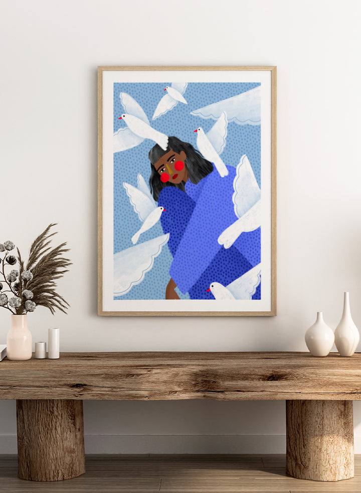 A framed art print featuring a stylized illustration of a woman with dark hair and red cheeks, wrapped in a blue patterned blanket, surrounded by white doves against a light blue dotted background. Peaceful Woman and Doves by Bea Müller art print featuring a woman in blue surrounded by white birds, layered textures, and soft modern color.