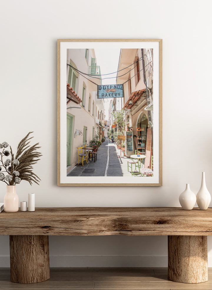  narrow street with a bakery sign in a European town. Charming Bakery Street by Henrike Schenk is a Greek street photography print featuring a Mediterranean bakery alley, pastel buildings, café seating, and travel wall art.