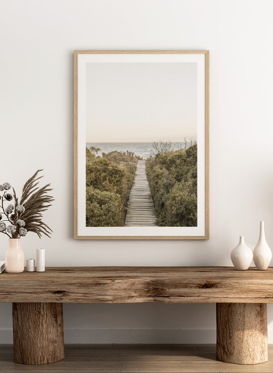A framed photographic art print depicting a wooden boardwalk leading from dense green beach vegetation to the ocean under a pale sky. Pathway to Tranquility by Shot by Clint coastal art print featuring a wooden boardwalk, wild greenery, distant palms, and a soft ocean horizon.