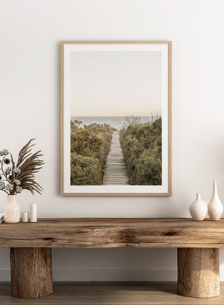 A framed photographic art print depicting a wooden boardwalk leading from dense green beach vegetation to the ocean under a pale sky. Pathway to Tranquility by Shot by Clint coastal art print featuring a wooden boardwalk, wild greenery, distant palms, and a soft ocean horizon.