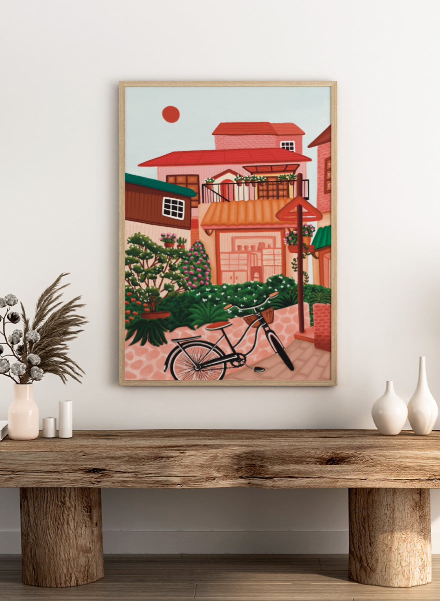 A colorful illustrated street scene with pink and peach buildings, green plants, flowers, a bicycle in the foreground, and a pale sky with a red sun. Shop Charming Village Street by Nidhi Kachhadiya, a colorful village wall art print featuring a bicycle, flower-filled homes, terracotta roofs, and warm pink and green tones.