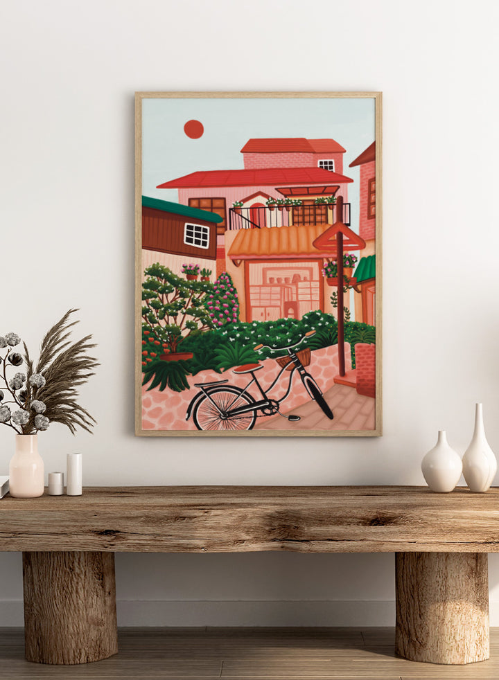 A colorful illustrated street scene with pink and peach buildings, green plants, flowers, a bicycle in the foreground, and a pale sky with a red sun. Shop Charming Village Street by Nidhi Kachhadiya, a colorful village wall art print featuring a bicycle, flower-filled homes, terracotta roofs, and warm pink and green tones.