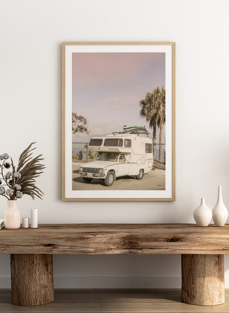 A vintage white camper truck parked outdoors near palm trees and a fence, with a surfboard strapped to its roof rack. Vintage Camper Parked by Shot by Clint travel photography print featuring a retro camper, surfboards, palm trees, and sun-faded coastal tones.