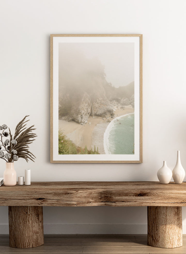 A framed photographic print of a misty coastal beach scene with a small waterfall, sandy shore, and pale blue-green ocean waves. Misty Coastal Haven by Shot by Clint coastal photography print featuring a secluded cove, rocky cliffs, pale turquoise water, and a soft veil of mist.