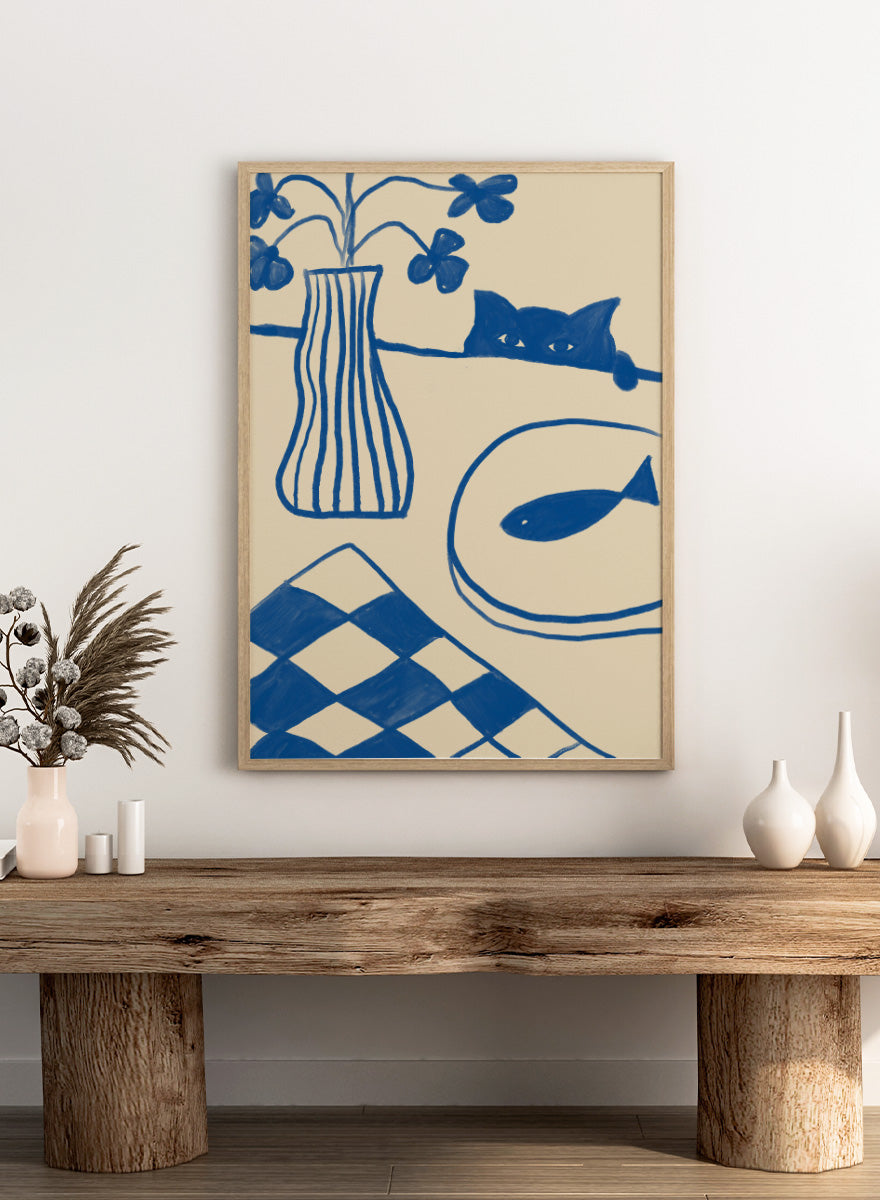 A framed art print featuring a hand-drawn style illustration in blue ink on a warm cream background. The design includes a striped vase with flowers, a cat peeking over a line, a fish on a plate, and a harlequin check pattern in the lower left corner. Blue Cat And Fish by The Art Concept is a playful cat art print featuring a curious blue cat, fish plate, striped vase, flowers, and checkered tablecloth.