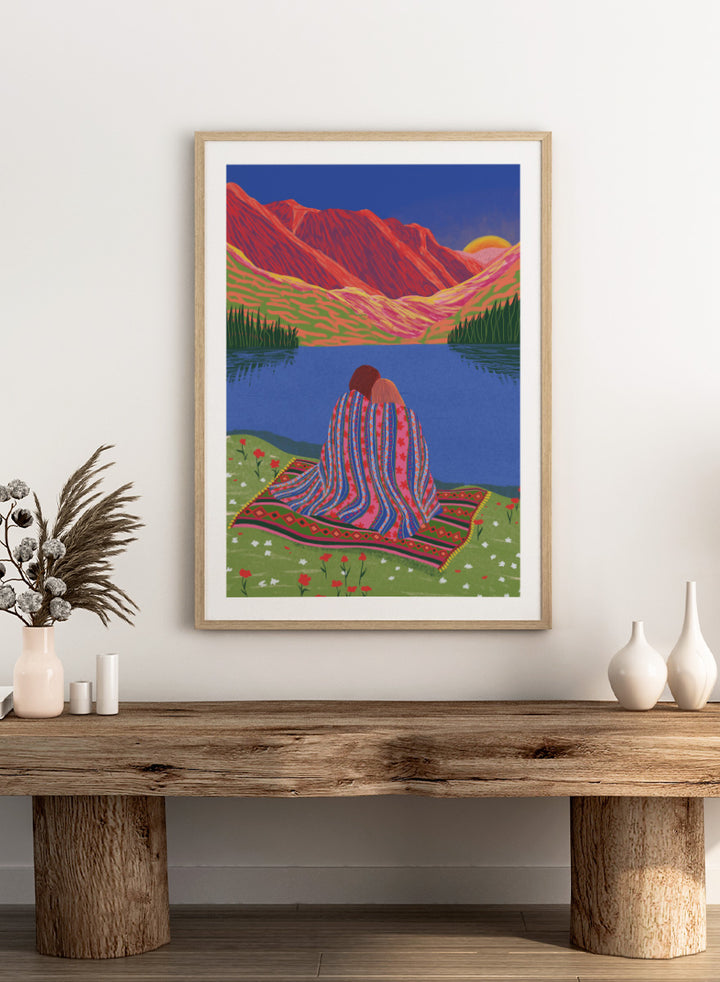 A framed art print featuring a stylized landscape illustration of two people sitting together on a patterned blanket by a blue lake, with red mountains and a yellow sun in the background. Cozy Lake Sunset art print by Ceyda Alasar features a lakeside embrace, vivid mountains, and a glowing sunset in rich, story-filled color.