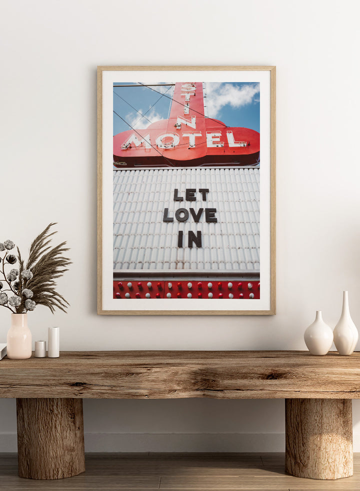 Vintage motel sign with 'Let Love In' message against a blue sky. Let Love In by Bethany Young features a vintage motel sign with red neon lettering against blue sky. Retro Americana photography wall art print.