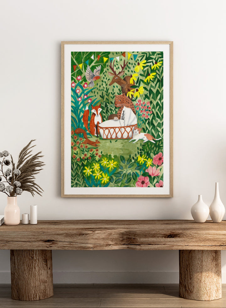 A framed art print featuring a whimsical forest illustration with animals gathered around a basket, surrounded by flowers and foliage. The print is shown with a white mat and a black frame against a white background. Welcome to the Forest by Caroline Bonne Müller woodland animal art print featuring a baby surrounded by a leopard, fox, deer, cat, flowers, and forest life.
