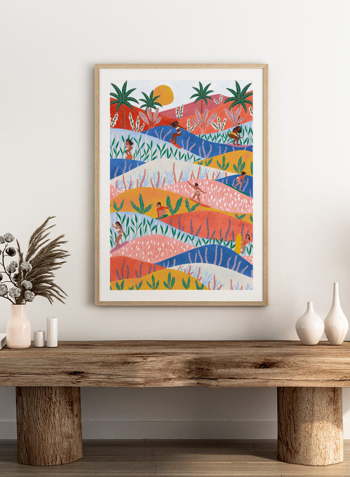 A framed art print featuring a colorful, stylized landscape illustration with rolling hills, palm trees, a sun, and small human figures in a folk-art style. Tropical Surf Girls by Ceyda Alasar surf art print featuring women riding waves through a colorful palm-filled landscape in bold tropical tones.