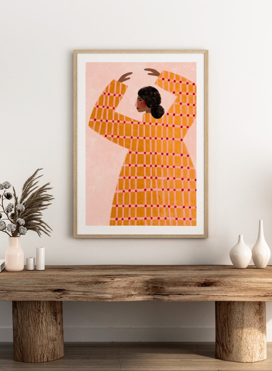 A framed art print featuring a stylized illustration of a woman in profile wearing a bright orange dress with a red grid pattern, set against a pale pink background. Dancing Woman in Orange by Bea Müller premium fine art print featuring a bold figure, warm orange tones, and a playful graphic composition.