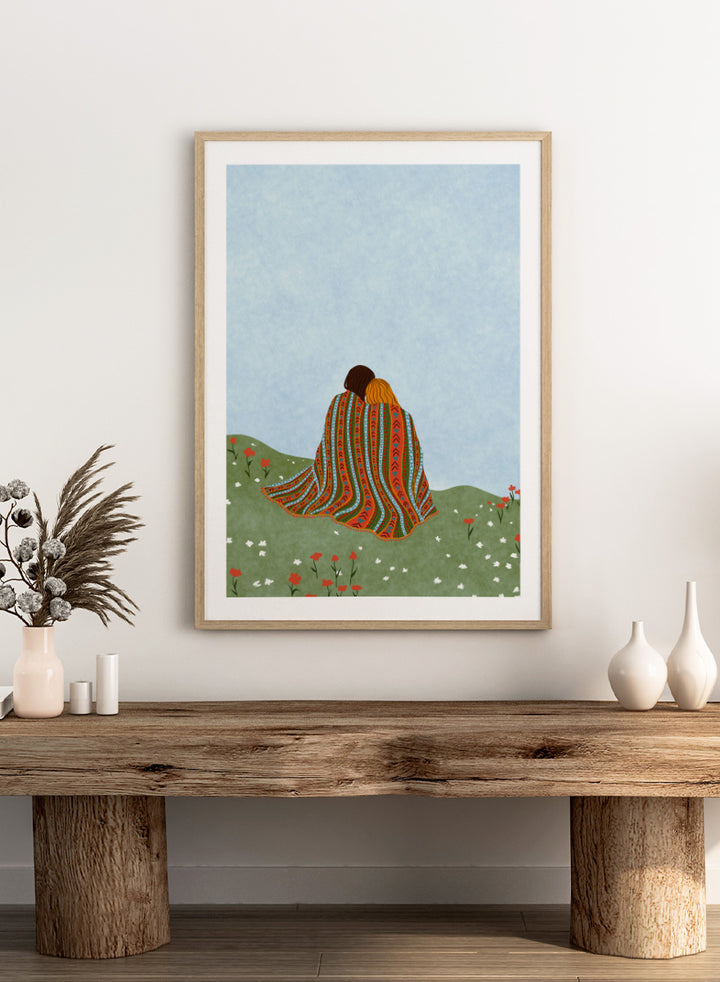A framed art print featuring an illustration of two people sitting on a grassy hill, wrapped in a vibrant, patterned blanket under a light blue sky. Cozy Hillside Embrace by Ceyda Alasar art print featuring two people wrapped in a patterned blanket among green hills and wildflowers.