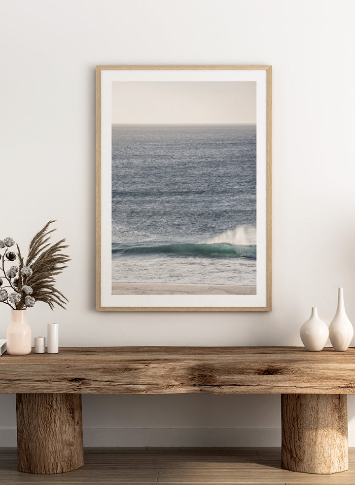 A framed photographic art print of an ocean scene, featuring a gentle wave breaking on a sandy beach under a pale sky. Tranquil Ocean Scene by Shot by Clint ocean photography print with a rolling wave, soft sea spray, and airy blue coastal tones.