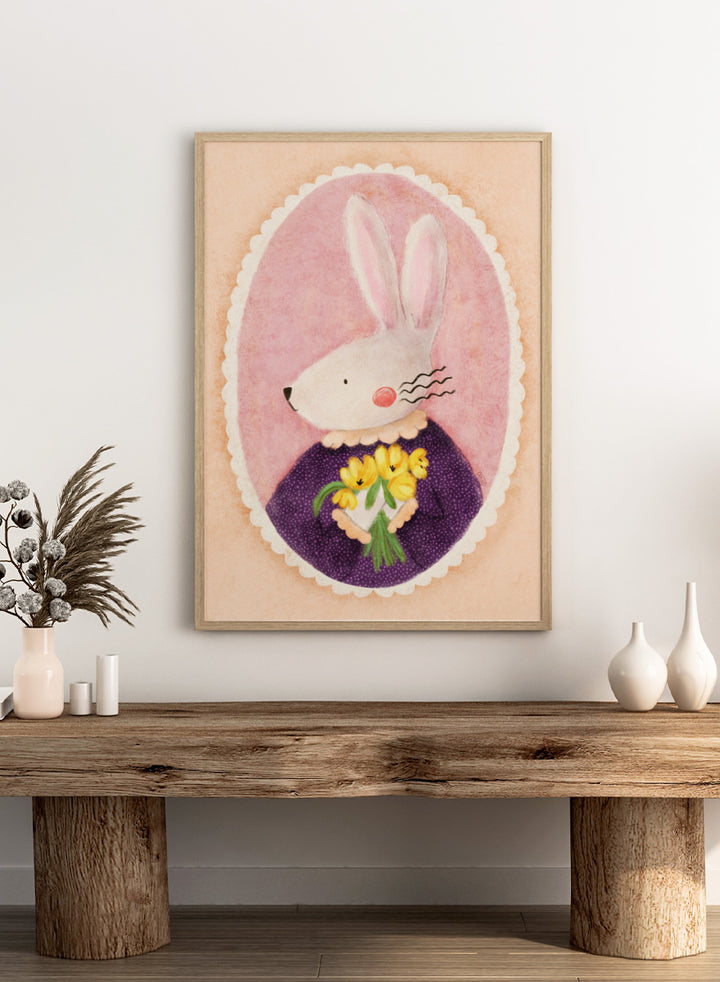 A framed art print featuring a whimsical illustration of a white rabbit in a purple dress holding yellow flowers, set against a pink oval background. Shop Charming Bunny With Tulips by Dirkie Nell, a playful animal portrait art print featuring a white bunny, yellow tulips, purple dress, and soft pink tones.