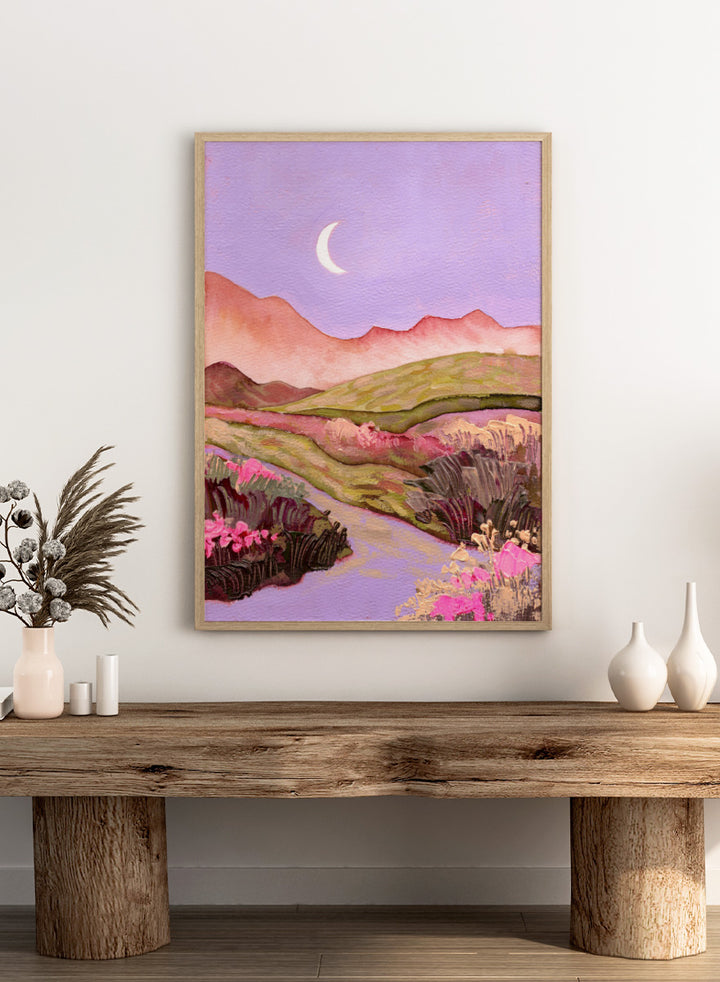 A landscape painting featuring rolling green hills, pink mountains, a purple sky with a crescent moon, and pink flowers along a winding path. Shop Moonlit Valley Path by Ekaterina Prisich, a colorful landscape art print featuring a crescent moon, winding path, rolling hills, and wildflowers.