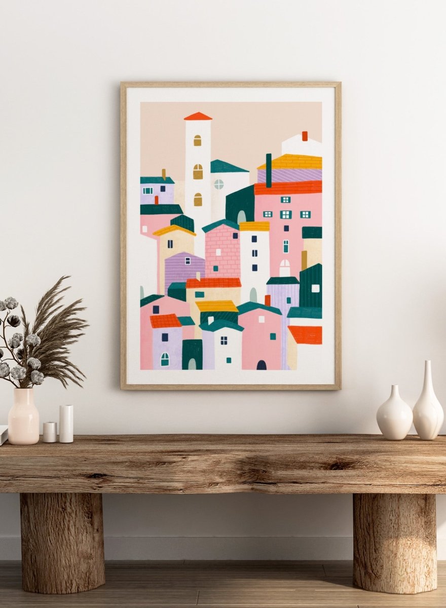 A framed minimalist art print depicting a cluster of stylized pastel houses and a church tower in pink, peach, teal, yellow, and cream tones. Charming Colorful Houses by Bea Müller premium framed fine art print featuring a playful town of pink, lavender, cream, and teal buildings.