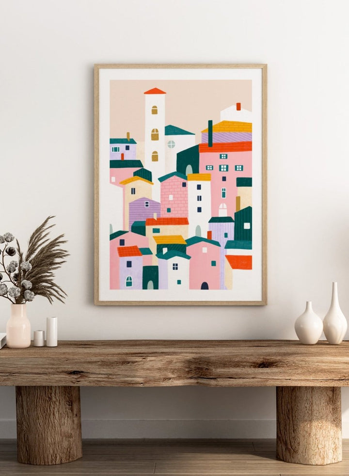 A framed minimalist art print depicting a cluster of stylized pastel houses and a church tower in pink, peach, teal, yellow, and cream tones. Charming Colorful Houses by Bea Müller premium framed fine art print featuring a playful town of pink, lavender, cream, and teal buildings.