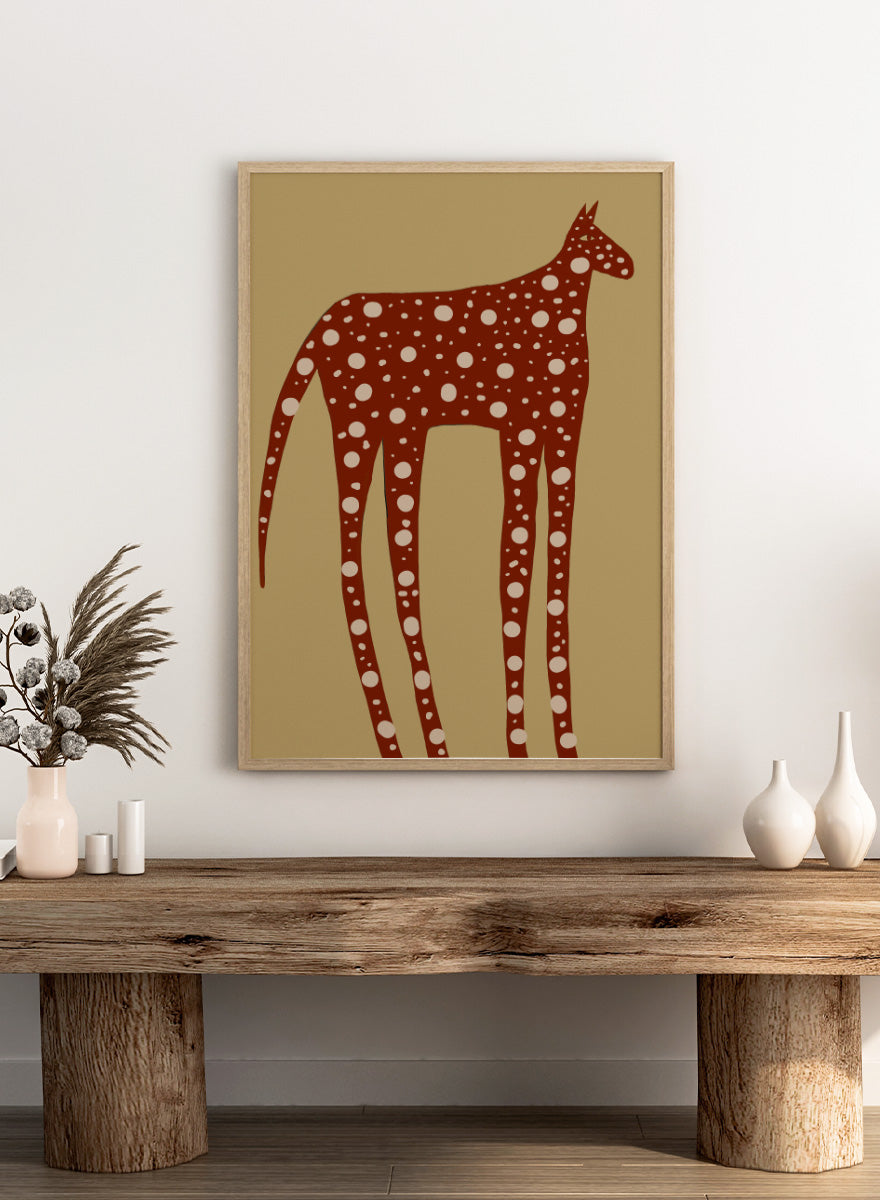 A stylized art print of a giraffe in reddish-brown with white spots, set against a tan background. Spotted Earthy Creature by The Art Concept animal art print featuring a long-legged rust-colored figure with cream spots on a muted tan background.