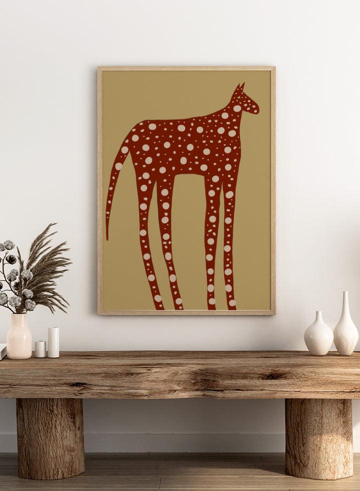 A stylized art print of a giraffe in reddish-brown with white spots, set against a tan background. Spotted Earthy Creature by The Art Concept animal art print featuring a long-legged rust-colored figure with cream spots on a muted tan background.