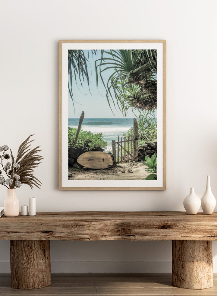 A framed beach scene art print featuring a wooden surfboard leaning against a rustic gate, with palm leaves framing a view of the ocean under a light blue sky. Coastal Serenity by Shot by Clint coastal art print featuring a surfboard, tropical plants, rustic gate, sandy path, and ocean waves.