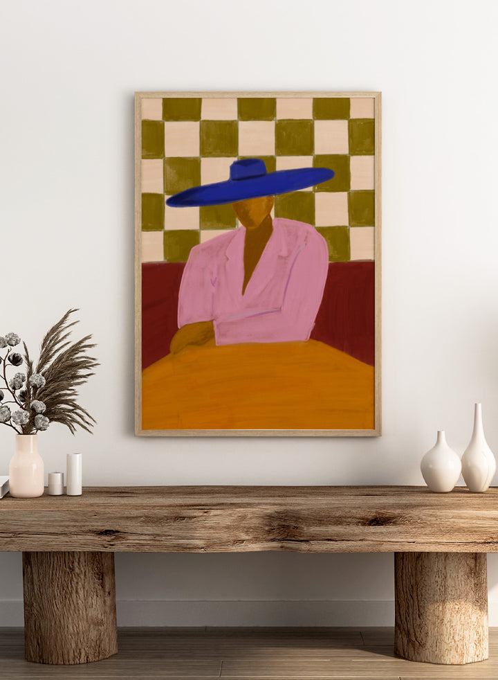 A stylized painting of a seated figure wearing a wide blue hat and pink top, with a red couch and a beige-and-olive checkered wall behind them. Blue Hat Chic by The Art Concept portrait art print featuring a cobalt blue hat, pink jacket, checkerboard backdrop, and bold modern color.