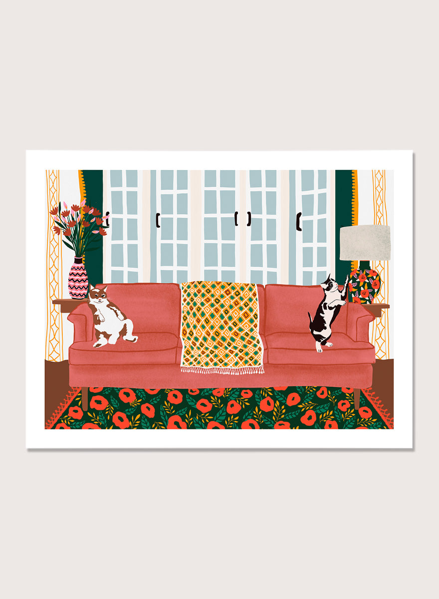 A framed art print depicting a cozy living room scene with a coral-pink sofa, two cats, a patterned throw blanket, and a floral rug. Playful Cats Living Room by Ceyda Alasar cat art print featuring two mischievous cats, a coral sofa, floral rug, patterned throw, and colorful decor.