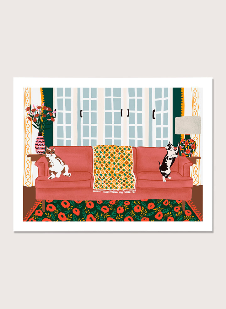A framed art print depicting a cozy living room scene with a coral-pink sofa, two cats, a patterned throw blanket, and a floral rug. Playful Cats Living Room by Ceyda Alasar cat art print featuring two mischievous cats, a coral sofa, floral rug, patterned throw, and colorful decor.