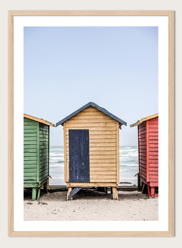 A framed photographic print of three colorful wooden beach huts on a sandy beach, with the central hut in natural wood and a dark blue door, flanked by green and red huts under a pale blue sky. Colorful Beach Huts by Shot by Clint coastal photography print featuring green, natural wood, and red huts with weathered texture and ocean views.