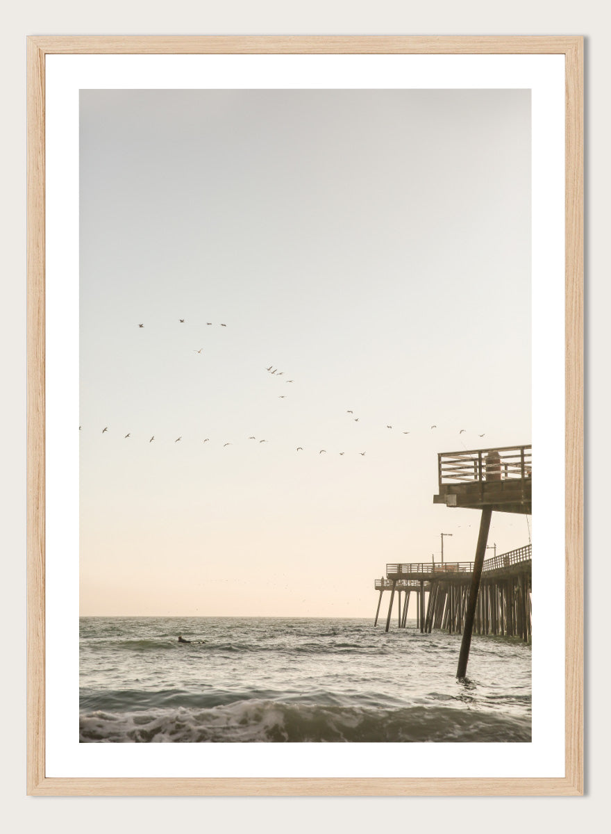 A framed photographic art print depicting a pier extending over a calm ocean under a pale sky, with birds flying across the scene. Seaside Serenity by Shot by Clint coastal photography print featuring ocean waves, a weathered pier, flying birds, and soft sunlit tones.