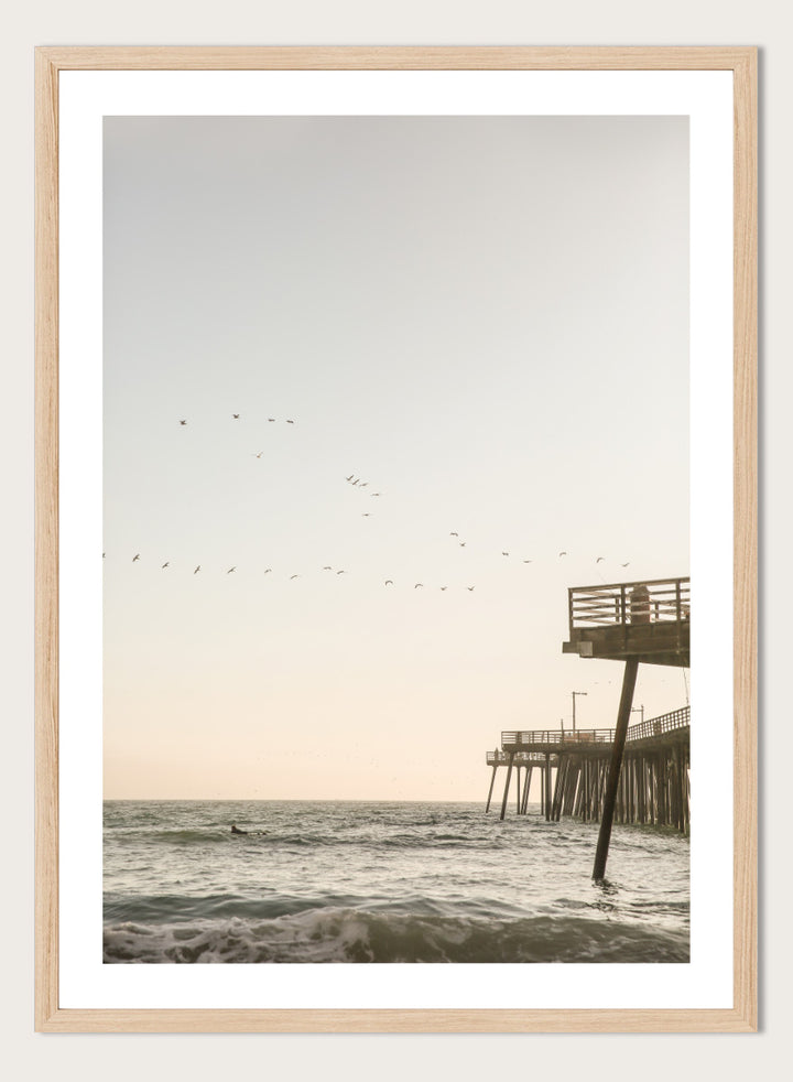 A framed photographic art print depicting a pier extending over a calm ocean under a pale sky, with birds flying across the scene. Seaside Serenity by Shot by Clint coastal photography print featuring ocean waves, a weathered pier, flying birds, and soft sunlit tones.