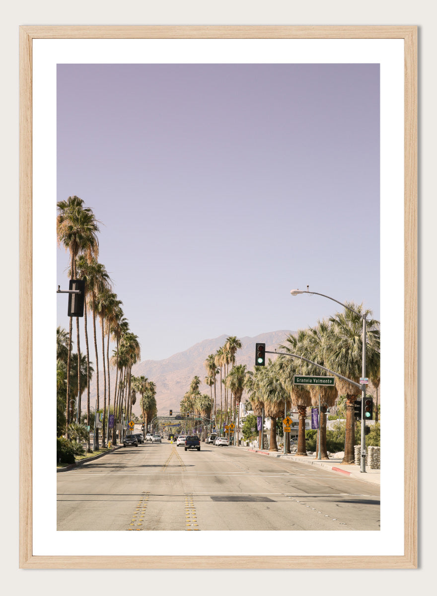 A framed art print featuring a scenic photograph of a palm-lined street in Palm Springs with mountains in the background. Palm-Lined Avenue by Shot by Clint travel photography print featuring a California road, tall palm trees, desert mountains, and soft sun-faded tones.