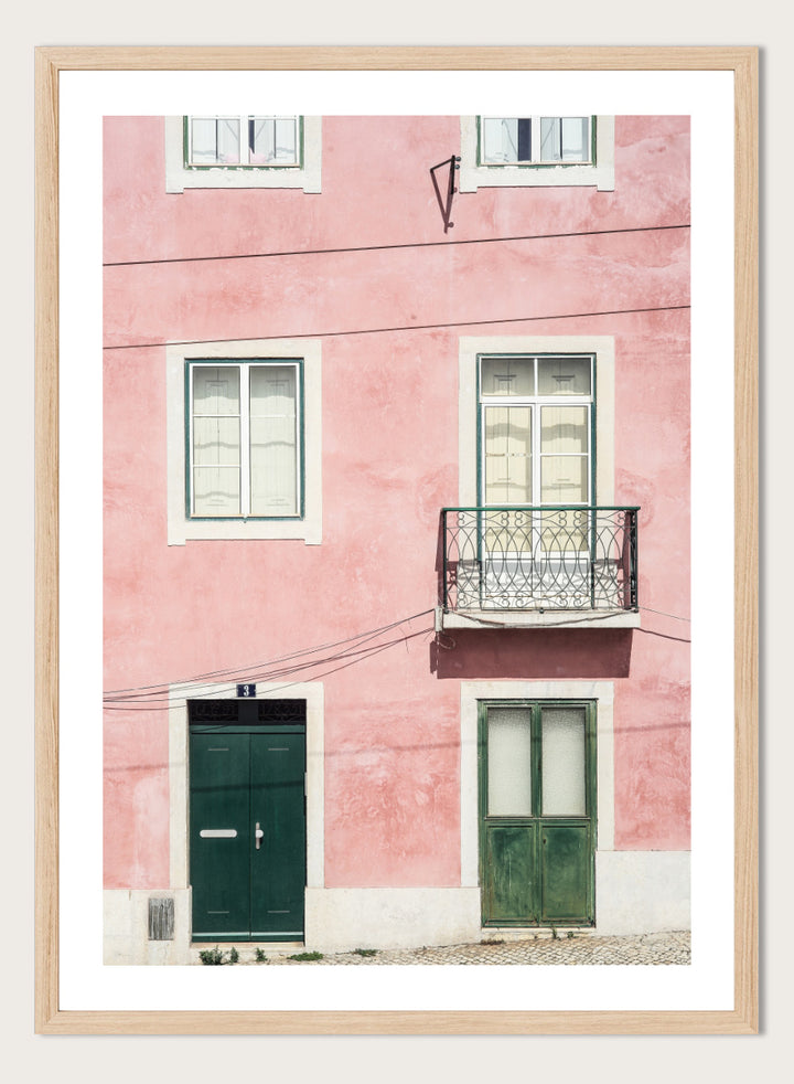 A framed art print featuring a photograph of a pink building facade with green doors, white-framed windows, and a small black metal balcony. Pink Façade Harmony by Shot by Clint architectural photography print featuring a pink exterior, green doors, white windows, and an iron balcony.